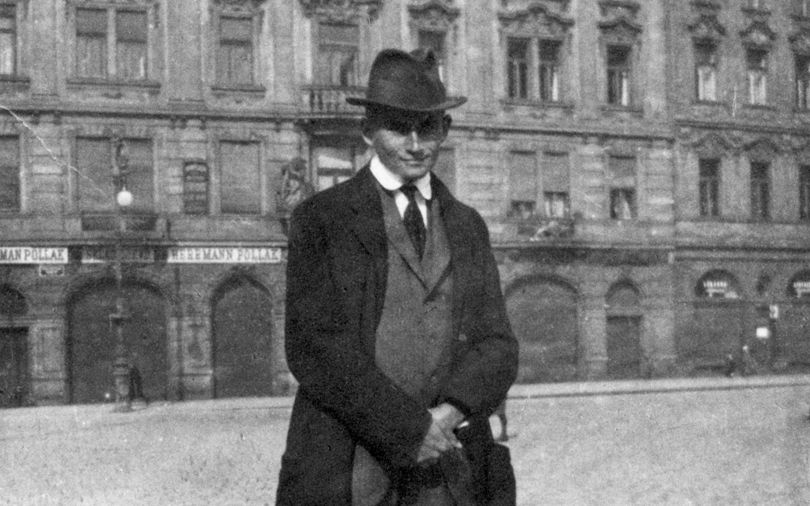 Black-and-white grainy photograph of Franz Kafka sating in fromt of a building wearing a long overcoat and hat.